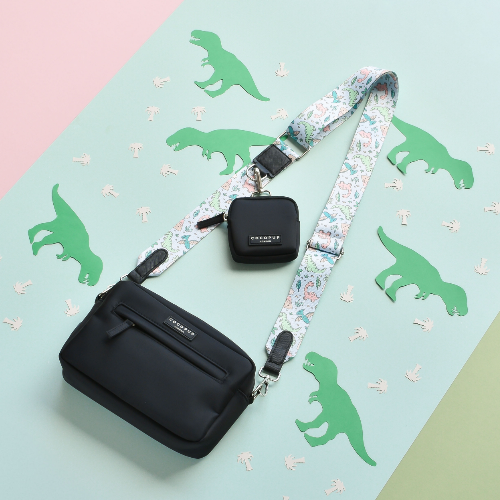 A stylish black crossbody bag featuring the Cocopup London Bag Strap - Dinopaws and a small matching pouch is placed on a pastel green and pink backdrop. Green dinosaur cutouts and white dino paw prints are arranged around the bag and pouch, creating a playful and whimsical scene.