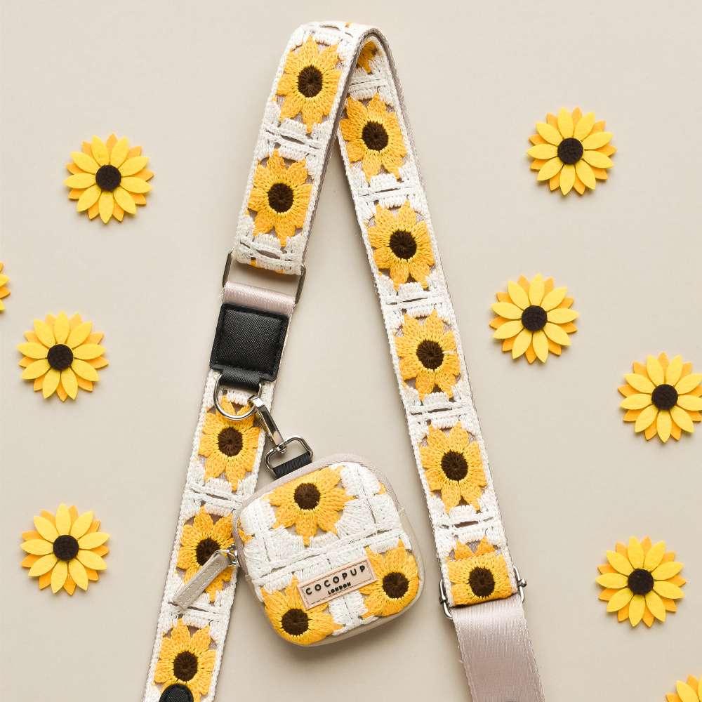 A small Dog Walking Bag and Cocopup London's Bag Strap - Sunflower Patch, both adorned with a sunflower pattern, are displayed on a beige background. Yellow and orange sunflowers are symmetrically arranged around the bag and strap, creating a balanced and visually appealing display that features crochet-style fabric.