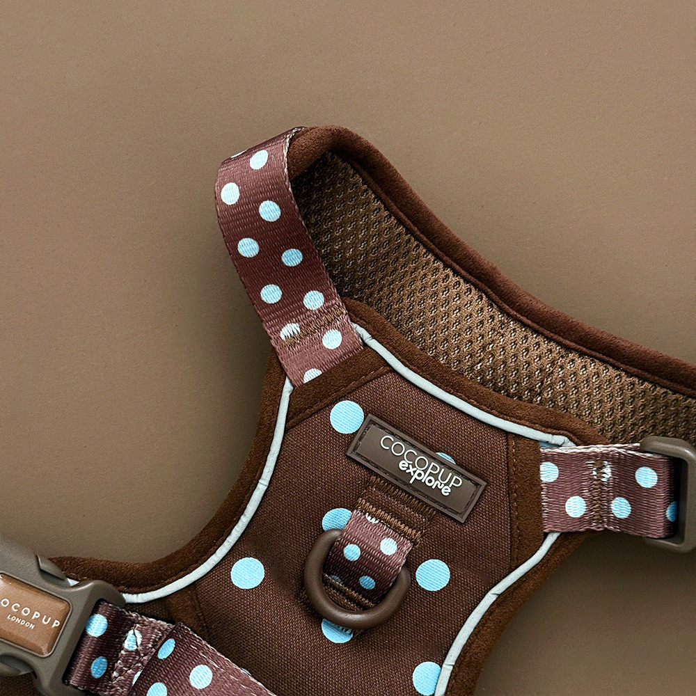 A close up of the Cocopup logo and d-ring on a brown dog harness with light blue polka dots, laid flat on a solid brown background. 