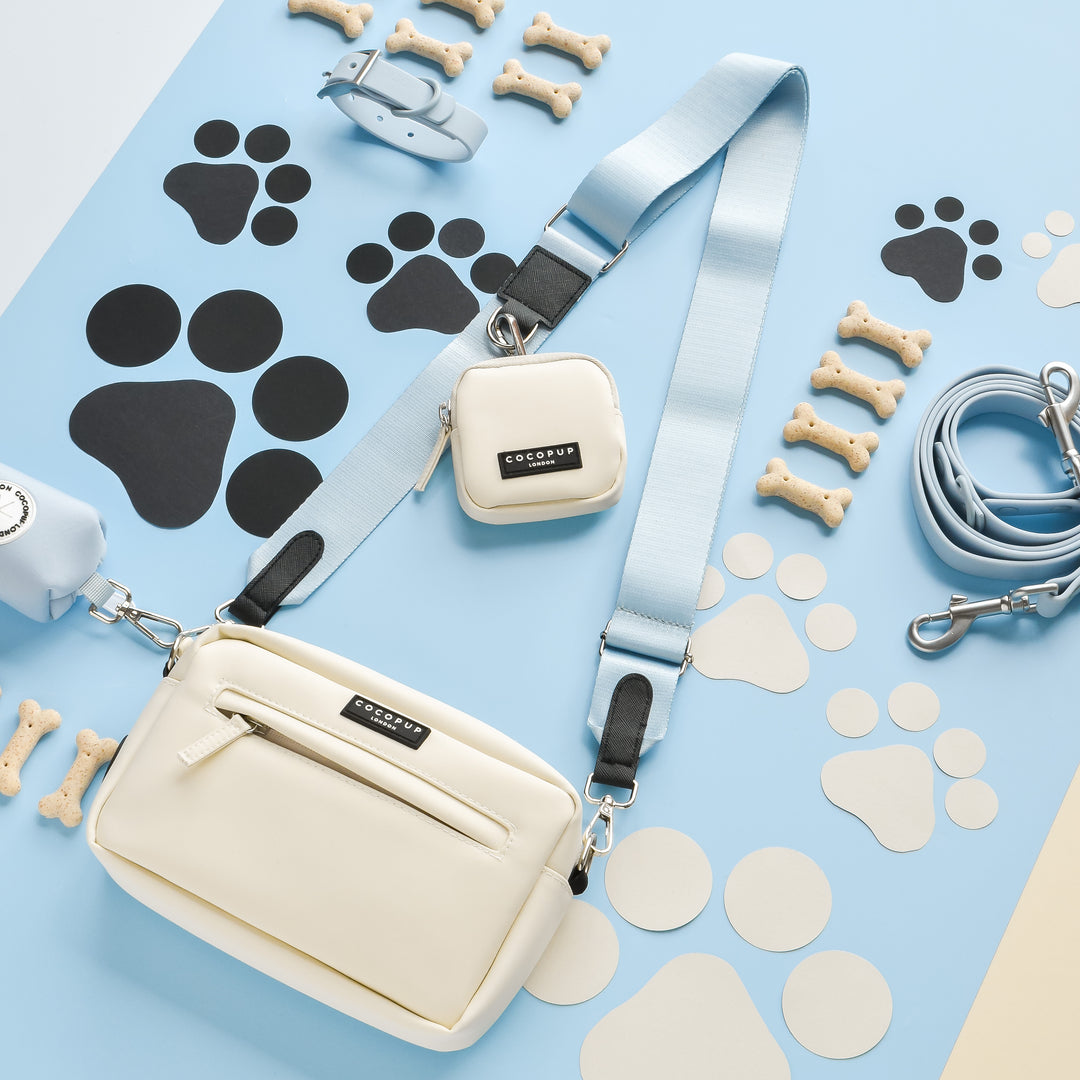 A flat lay image showcases a white Cocopup London Dog Walking Bag with a light blue NAKD Bag Strap - Bubblegum and a matching small pouch. Surrounding the bag are dog accessories, including a blue collar, leash, and bone-shaped treats. Black and white paw print cutouts adorn the background.