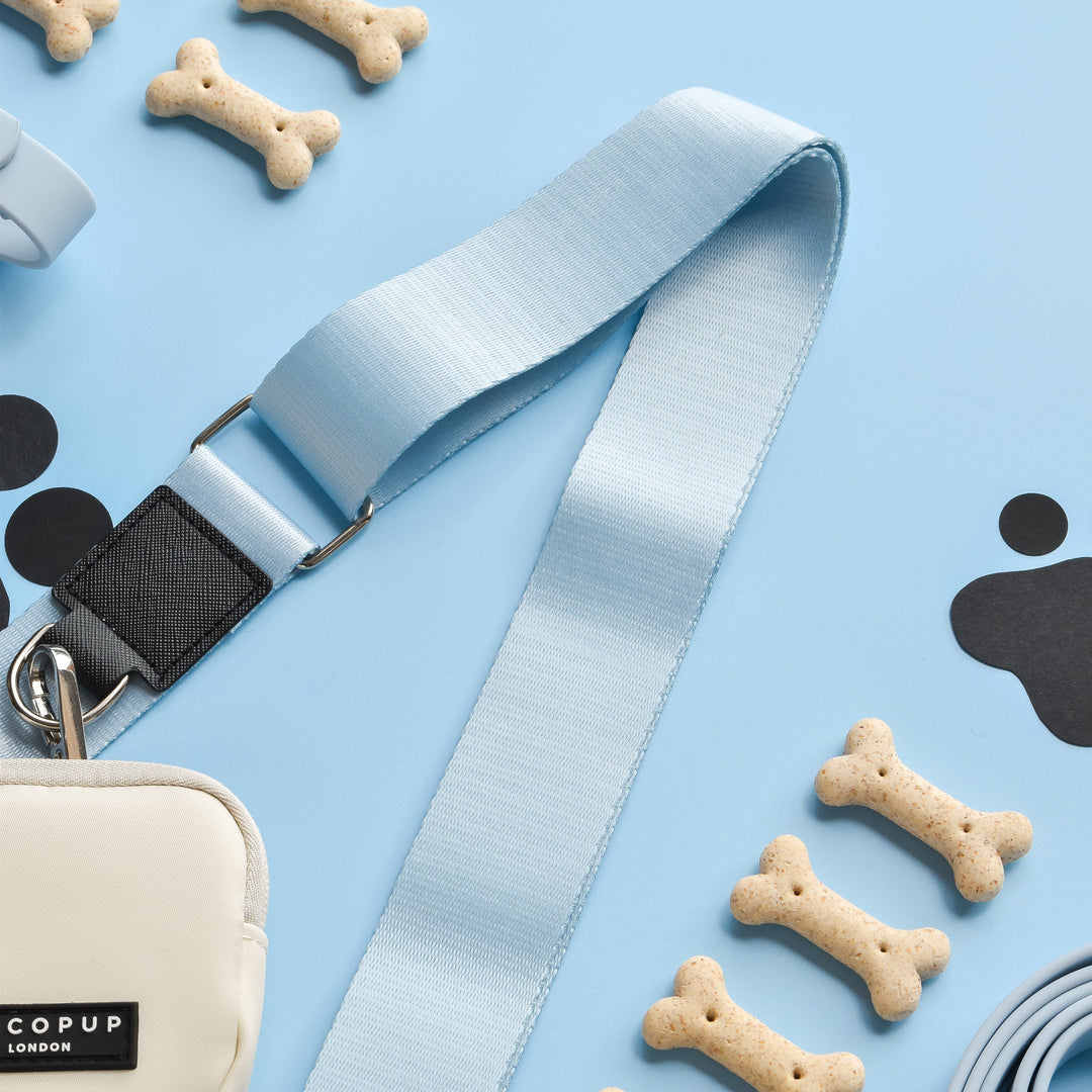 A flat lay of various dog accessories on a light blue background, featuring a light blue polyester leash, a small white Cocopup London Dog Walking Bag with a label, several dog treats shaped like bones, and black paw print cutouts. The bag is paired with the NAKD Bag Strap in Bubblegum.