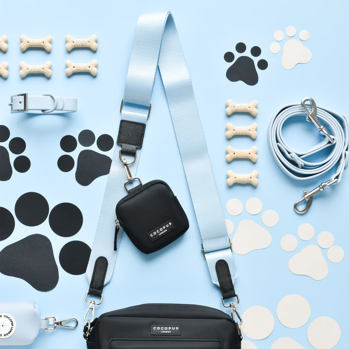 A light blue dog leash and collar, a black treat pouch, a Cocopup London Dog Walking Bag with a NAKD Bag Strap - Bubblegum in blue, and various dog bone treats are arranged neatly on a light blue background. Black and white paw print cutouts from the NAKD Bubblegum collection are scattered decoratively around the items.