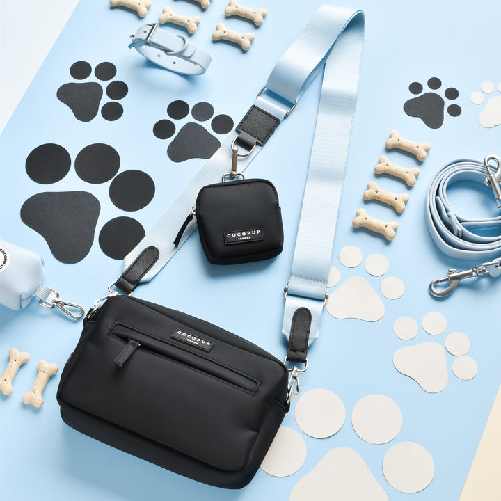 A stylish black Cocopup London Dog Walking Bag with a Bubblegum NAKD Bag Strap, a small matching black pouch, a blue collar, a blue leash, and various dog paw print shapes and bone-shaped treats arranged neatly on a light blue surface. Accessories suggest a pet theme.