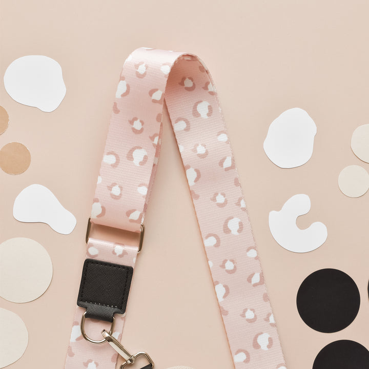 The Bag Strap - Nude Chic Leopard from Cocopup London, featuring a pink lanyard with a white abstract pattern, a black attachment, and a metal clip, is laid out flat on a beige background. Various white, black, and tan circular paper shapes are scattered around it, enhancing its stylish nude chic leopard vibe.