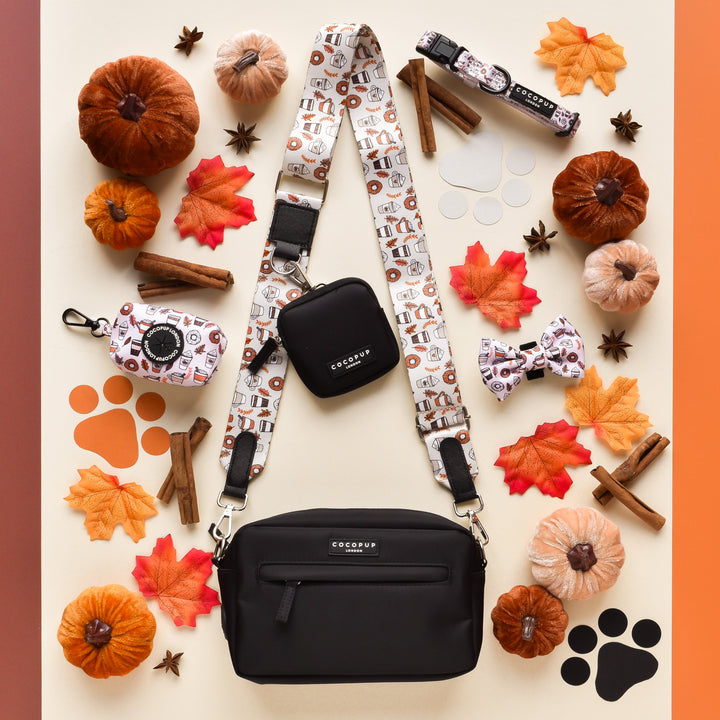 A flat lay of dog accessories from Cocopup London arranged on an autumn-colored background. Items include a black crossbody dog walking bag, the Bag Strap - Autumn Puppuccino, a poop bag holder, a bow tie, and a leash adorned with dog-themed patterns. The ensemble is surrounded by mini pumpkins, autumn leaves, cinnamon sticks, and paw prints—perfect for an Autumn Puppuccino outing.