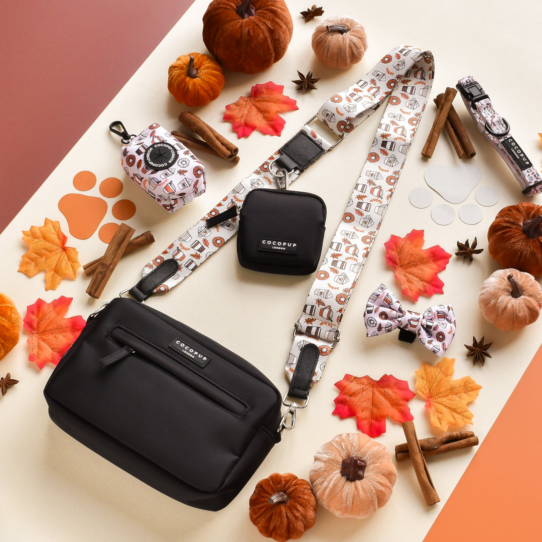 A black crossbody bag with the Cocopup London "Bag Strap - Autumn Puppuccino" featuring a fall-themed pattern is surrounded by small pumpkins, pinecones, leaves, and cinnamon sticks. Perfect as a dog walking bag, it includes matching accessories like a small pouch, a bow tie, and a mini pop case.
