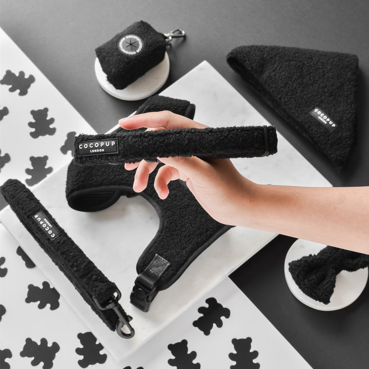 A hand holds a black fluffy dog accessory labeled "Cocopup London." The photo also showcases matching black dog items from the Teddy Baloo Adjustable Neck Harness, Lead & Collar Bundle. These include an adjustable neck harness, leash, poop bag holder, and bandana made from neoprene material against a black and white background with paw print designs.