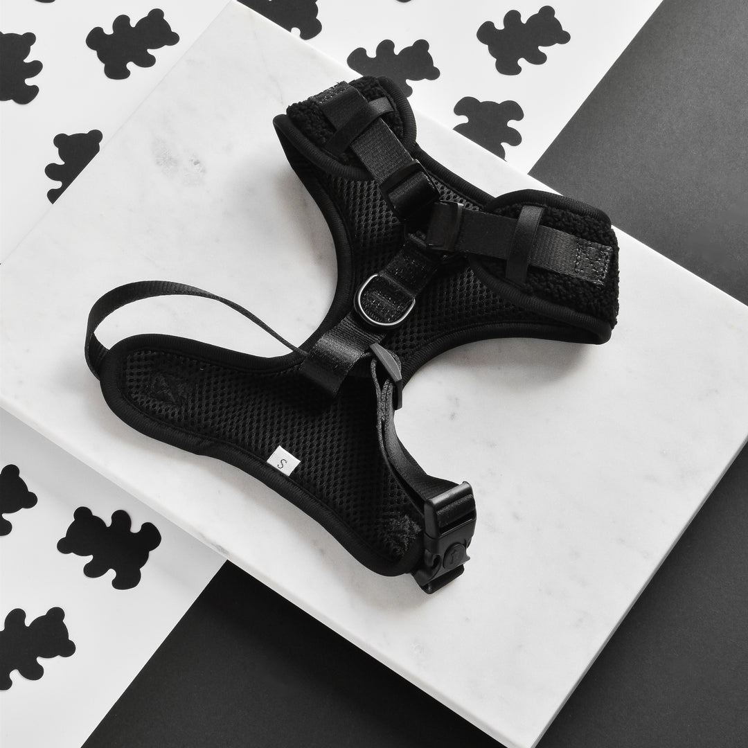 The Teddy Baloo Adjustable Neck Harness, Lead & Collar Bundle by Cocopup London, a black mesh dog harness made with neoprene material, featuring adjustable straps, a metal D-ring, and plastic buckles, is displayed on a white marble surface. The background showcases a monochrome pattern of bear silhouettes on a black and white backdrop.