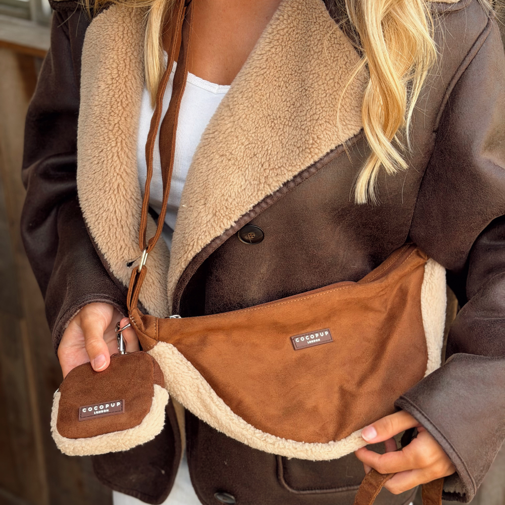 A person wearing a brown faux-SUEDE shearling bag with a matching small pouch, shown in a close-up lifestyle shot. The bag features a warm brown exterior and a beige sherpa lining, with a small brown brand patch reading 'COCOPUP LONDON'.