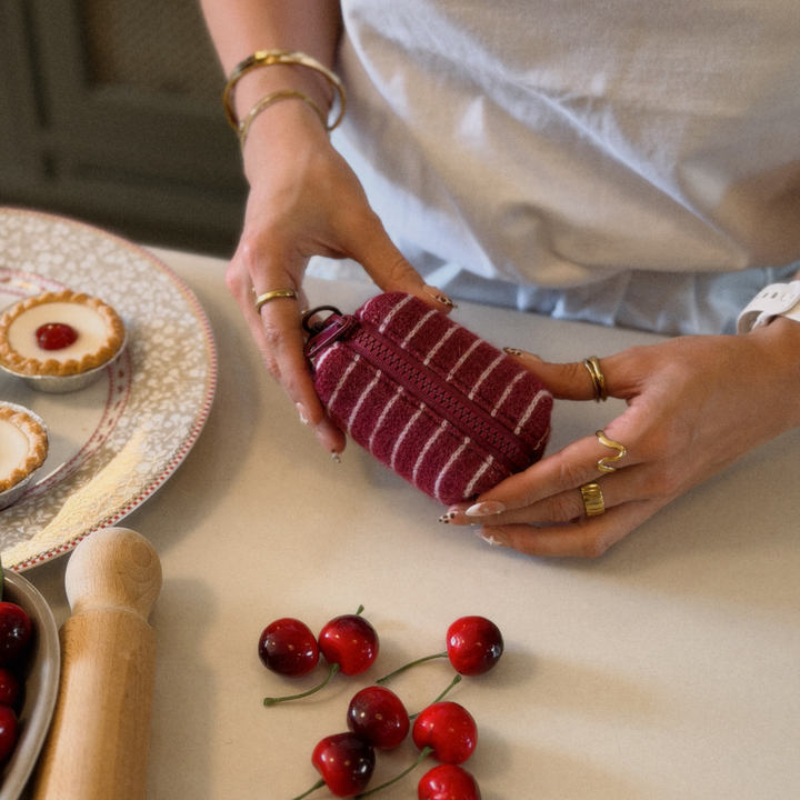 A person holds a small striped zippered poop bag holder in a kitchen setting, with cherries, a rolling pin, and tart pies visible on the counter.