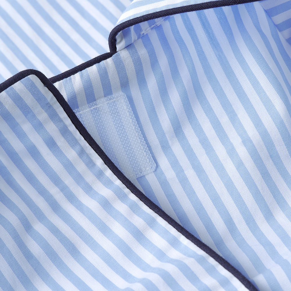Close-up of a light blue and white striped pajama top showing the collar, dark navy piping, and a hook-and-loop closure tab.