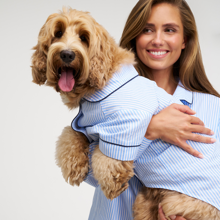 A woman holding a fluffy golden dog wearing a light blue and white striped pajama shirt with dark piping, against a clean light gray background.