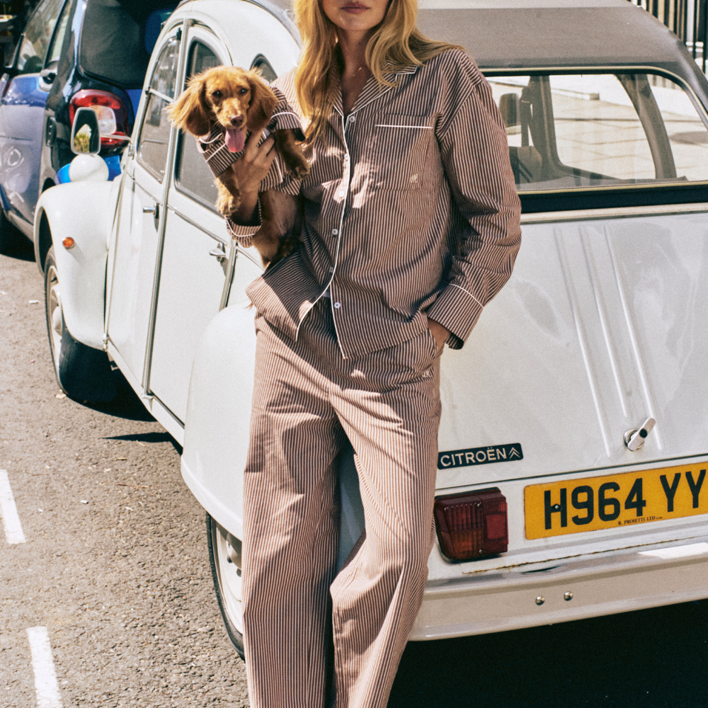A woman wearing a brown and beige pinstriped pajama set stands next to a vintage white Citroëen car, holding a small brown dog.